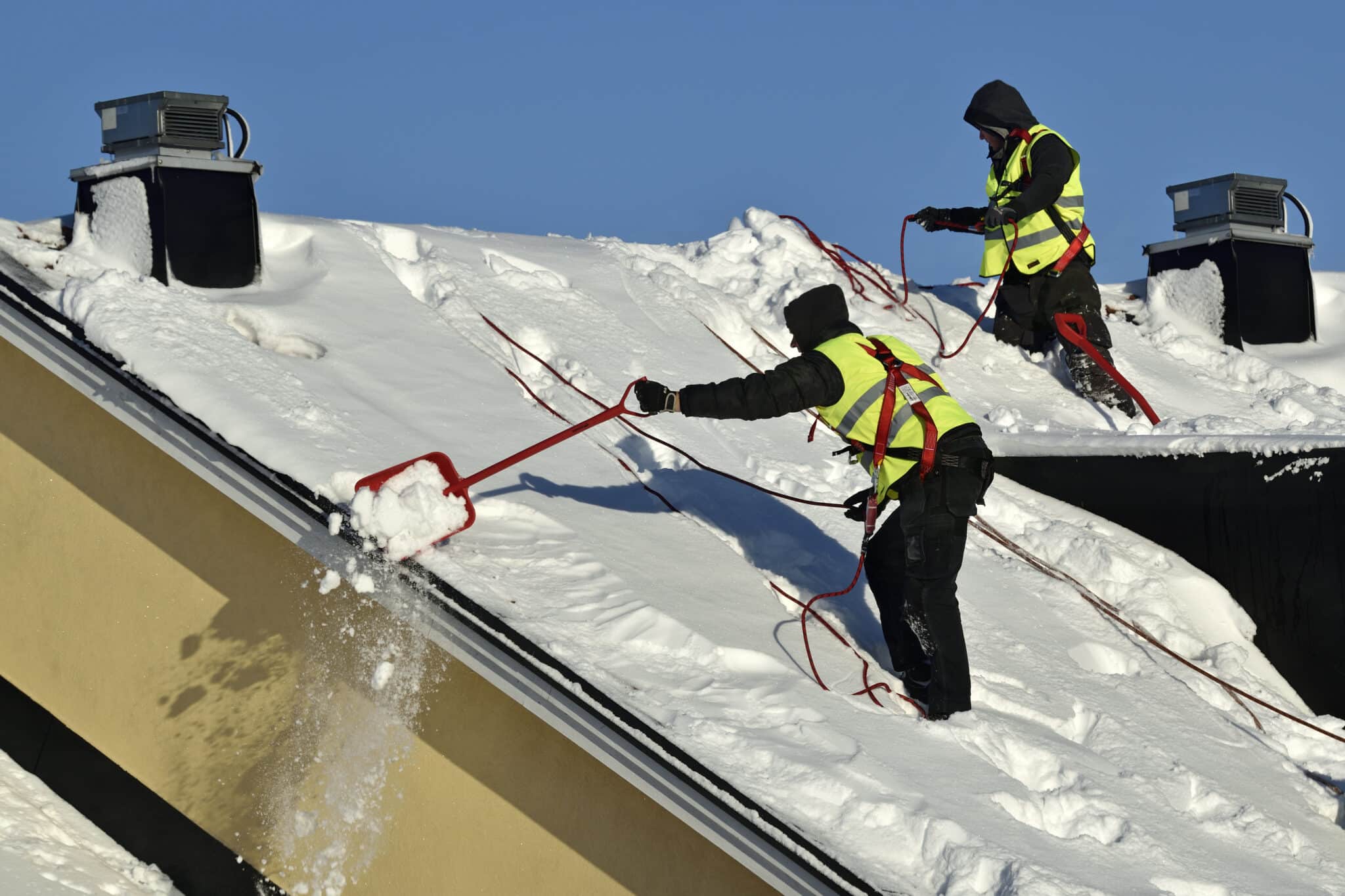 Workers with snow shovels carry out winter cleaning of roof of building from snow and ice
